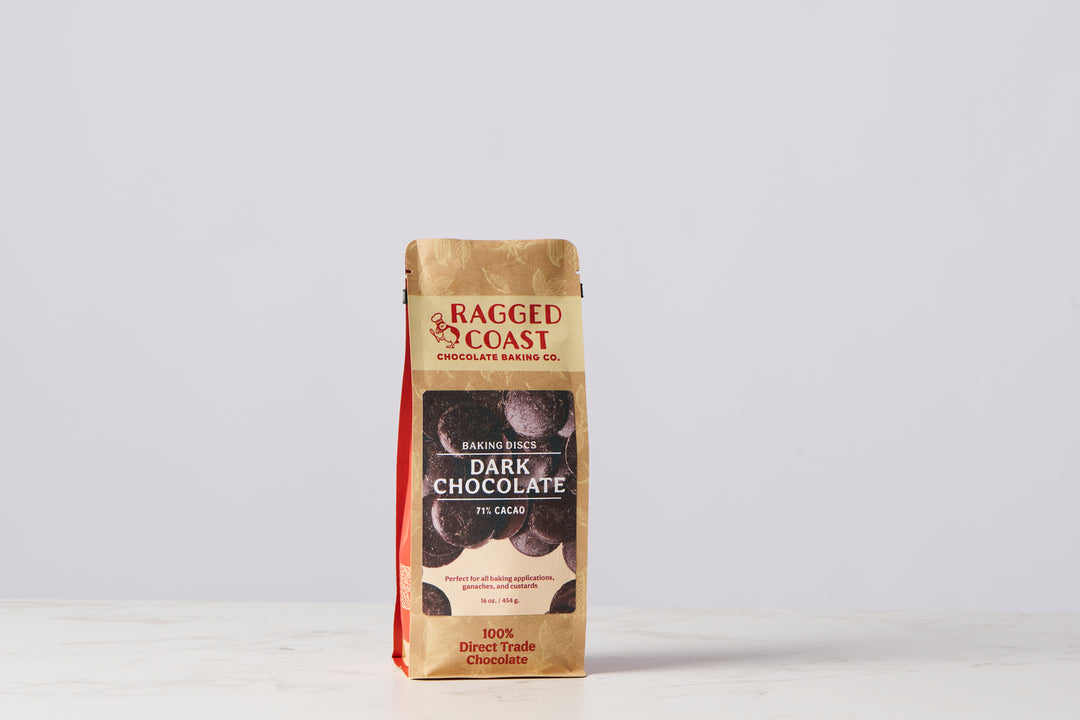 A brown bag of Ragged Coast Chocolates Bittersweet Chocolate Baking Discs, labeled "71% Cacao" and "100% Direct Trade Chocolate," is standing on a white surface against a plain background, highlighting its commitment to directly traded cacao and being non-GMO certified.