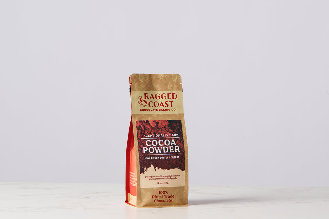 A bag of Ragged Coast Chocolates’ Exceptionally Dark Cocoa Powder, High Cocoa Butter Content - 12 ounces, distinctly labeled "Exceptionally Dark," "100% Direct Trade Chocolate," and "Dutch-processed," rests elegantly on a light marble surface with a plain background.