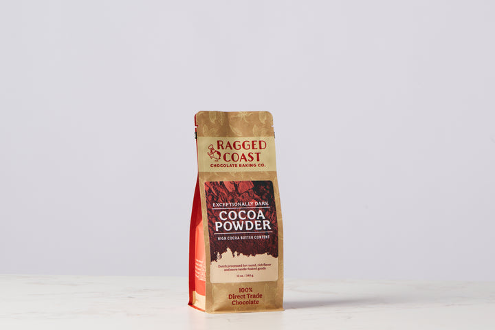 A bag of Ragged Coast Chocolates’ Exceptionally Dark Cocoa Powder, High Cocoa Butter Content - 12 ounces, distinctly labeled "Exceptionally Dark," "100% Direct Trade Chocolate," and "Dutch-processed," rests elegantly on a light marble surface with a plain background.