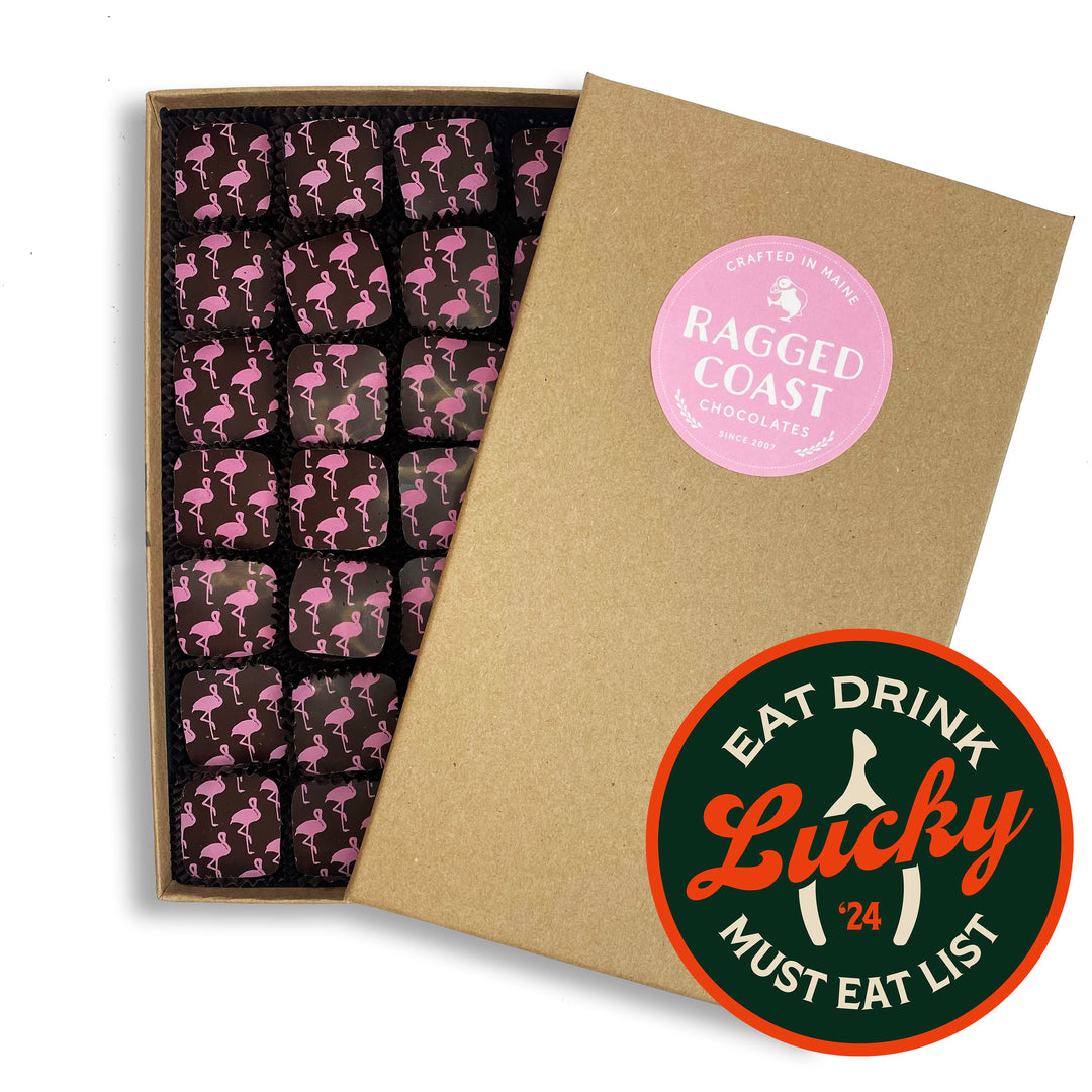 A box of Ragged Coast Chocolates featuring East Side Flamingo Truffles with pink flamingo designs on top, partially open. A green and red "Eat Drink Lucky '24 Must Eat List" sticker is placed on the bottom right corner of the box. The bittersweet chocolate pairs perfectly with a luscious white chocolate ganache filling.
