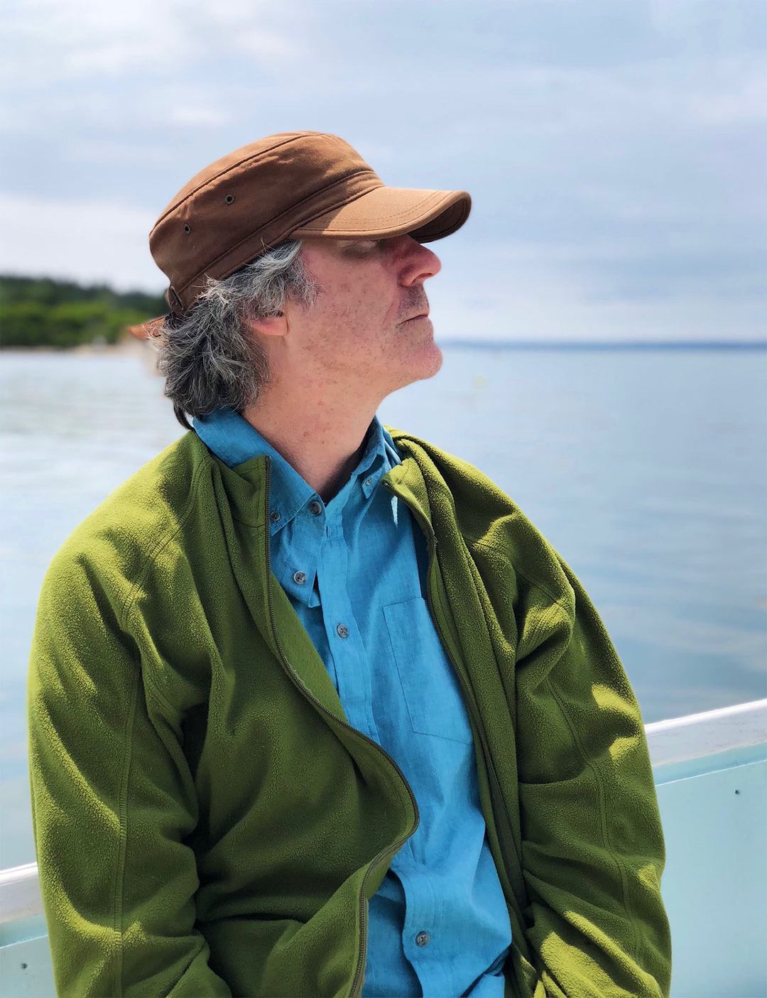 Man in a hat and green jacket looking contemplatively towards the water on a cloudy day.