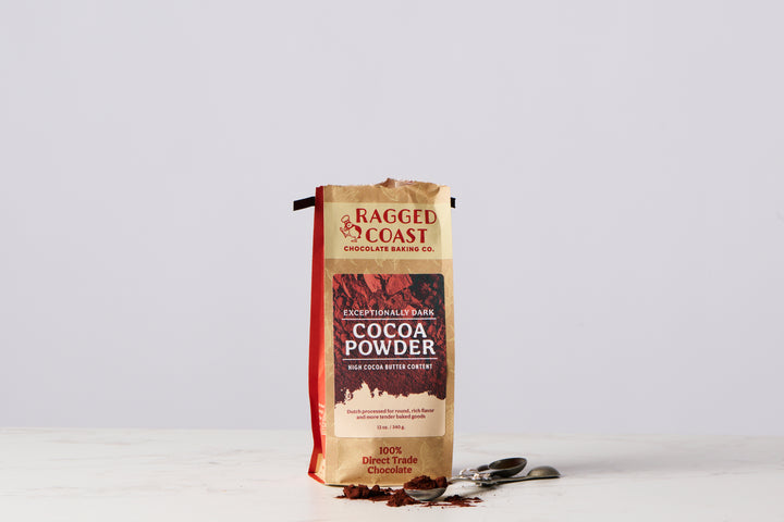 A standing package of Ragged Coast Chocolates' Exceptionally Dark Cocoa Powder, High Cocoa Butter Content - 12 ounces, is on a white surface, accompanied by a few scoops of Dutch-processed cocoa and three measuring spoons.