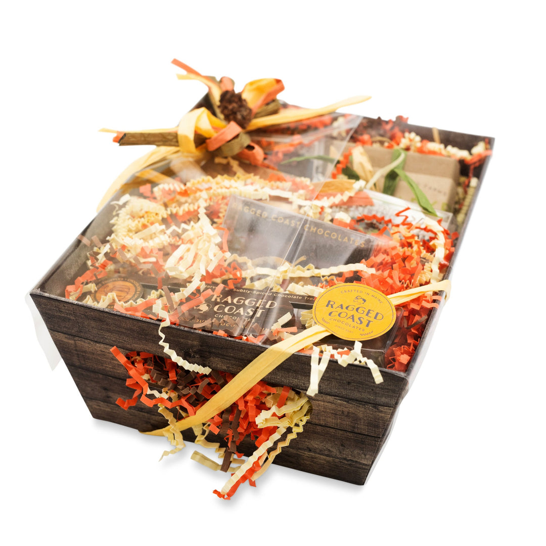 A gift basket from Ragged Coast Chocolates, featuring their Chocolate Holiday Subscription and assorted food items, beautifully adorned with ribbons and decorative shredded paper, all elegantly presented in a charming wooden box.