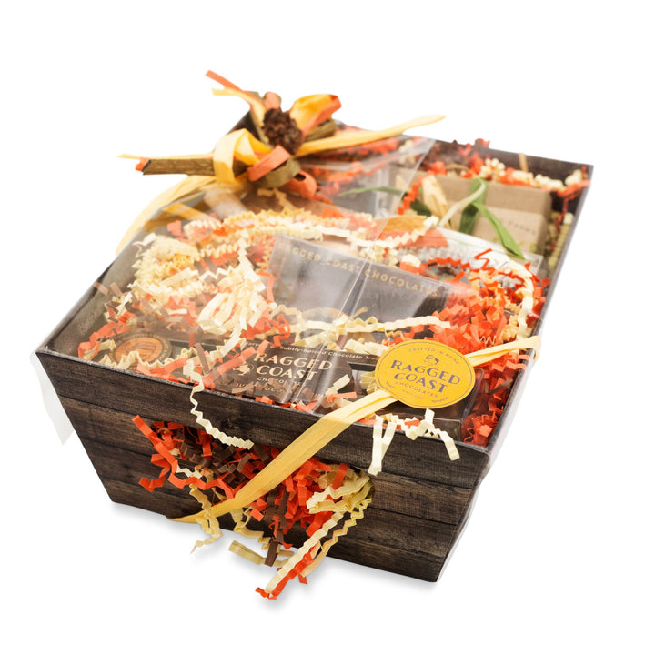 A gift basket from Ragged Coast Chocolates, featuring their Chocolate Holiday Subscription and assorted food items, beautifully adorned with ribbons and decorative shredded paper, all elegantly presented in a charming wooden box.