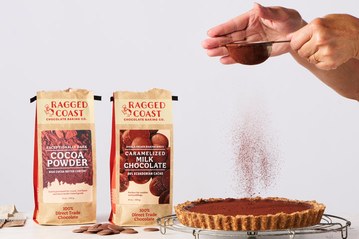 Hands sprinkling exceptionally dark cocoa powder over a chocolate pie. Two packages labeled "Ragged Coast Chocolates Exceptionally Dark Cocoa Powder, High Cocoa Butter Content - 12 ounces" and "Ragged Coast Caramelized Milk Chocolate" are standing in the background.
