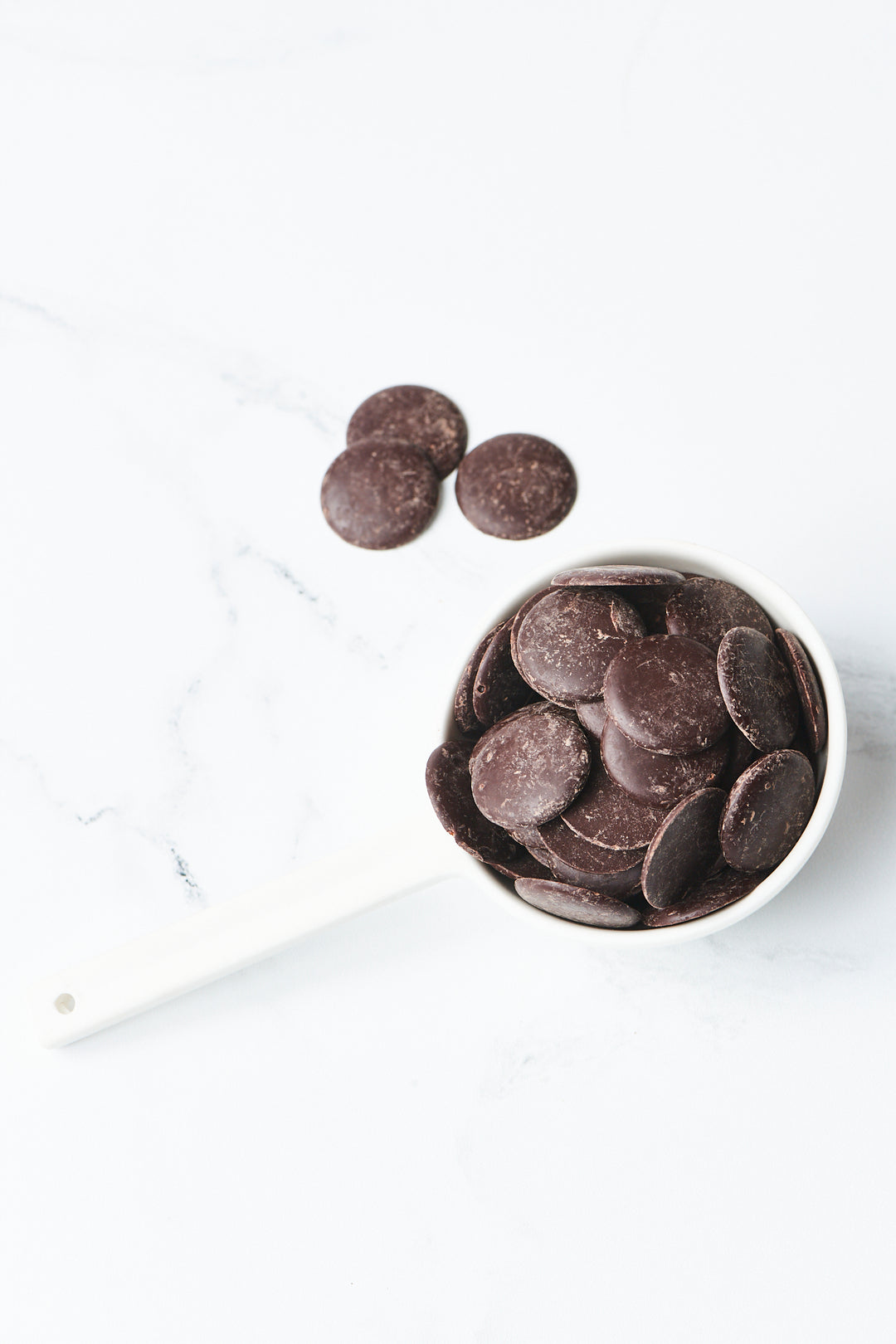 A white measuring cup filled with Ragged Coast Chocolates' Bittersweet Chocolate Baking Discs, 71% Cacao - 16 ounces is placed on a white marble surface. Additional non-GMO certified dark chocolate disks are scattered next to the cup.
