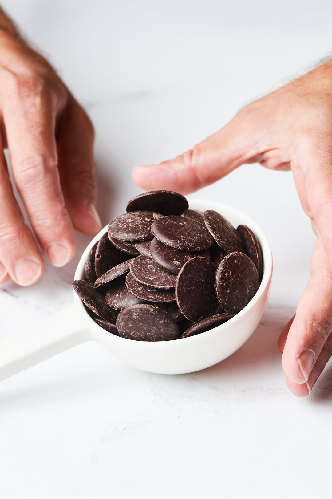 Hands are carefully holding and positioning a white bowl filled with Ragged Coast Chocolates' Bittersweet Chocolate Baking Discs, 71% Cacao - 16 ounces, on a light surface.