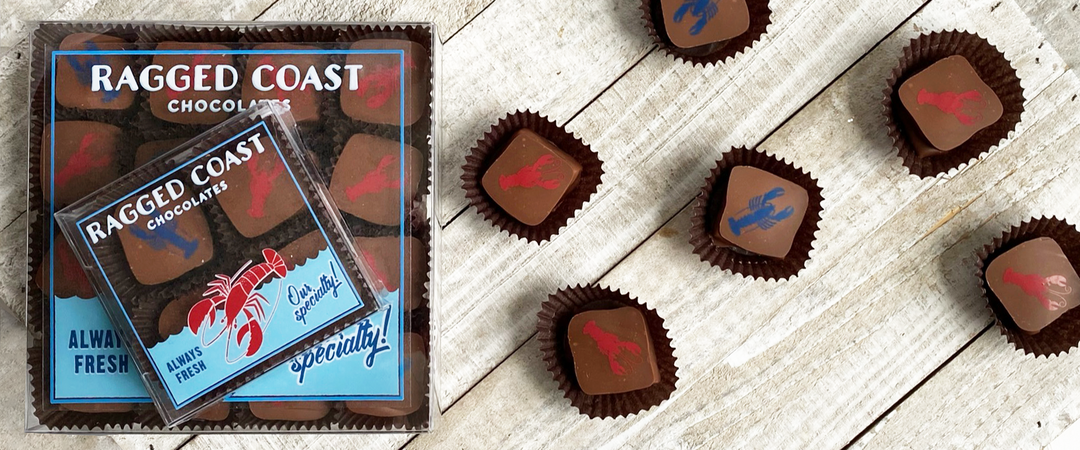 Image of a box of Ragged Coast Chocolates with some chocolates laid out on a wooden surface. The chocolates have red and blue lobster designs on them.