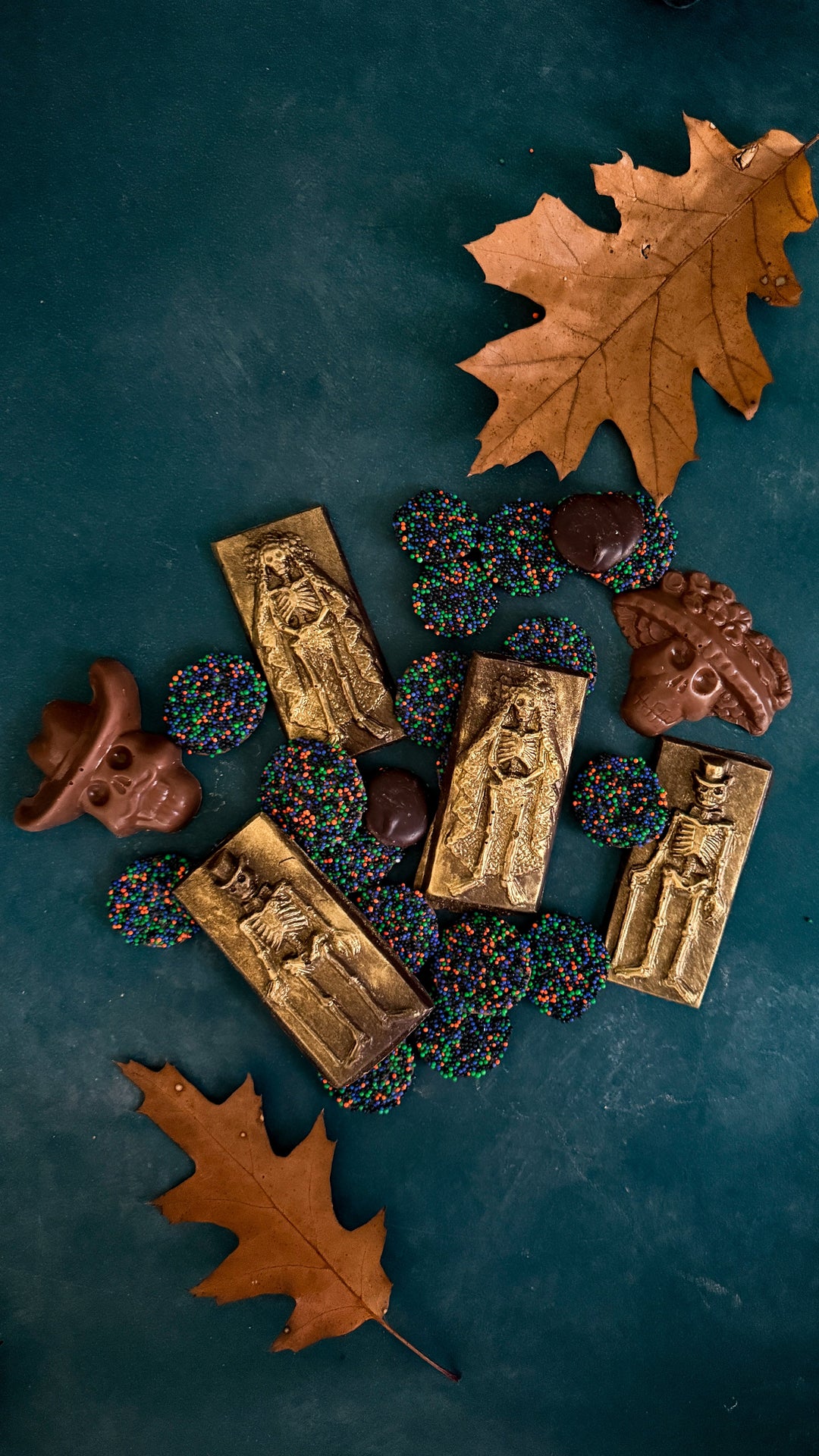 Gold-dusted chocolate skeleton bars and skull shapes surrounded by chocolate-coated sprinkles and two fallen oak leaves on a teal background.