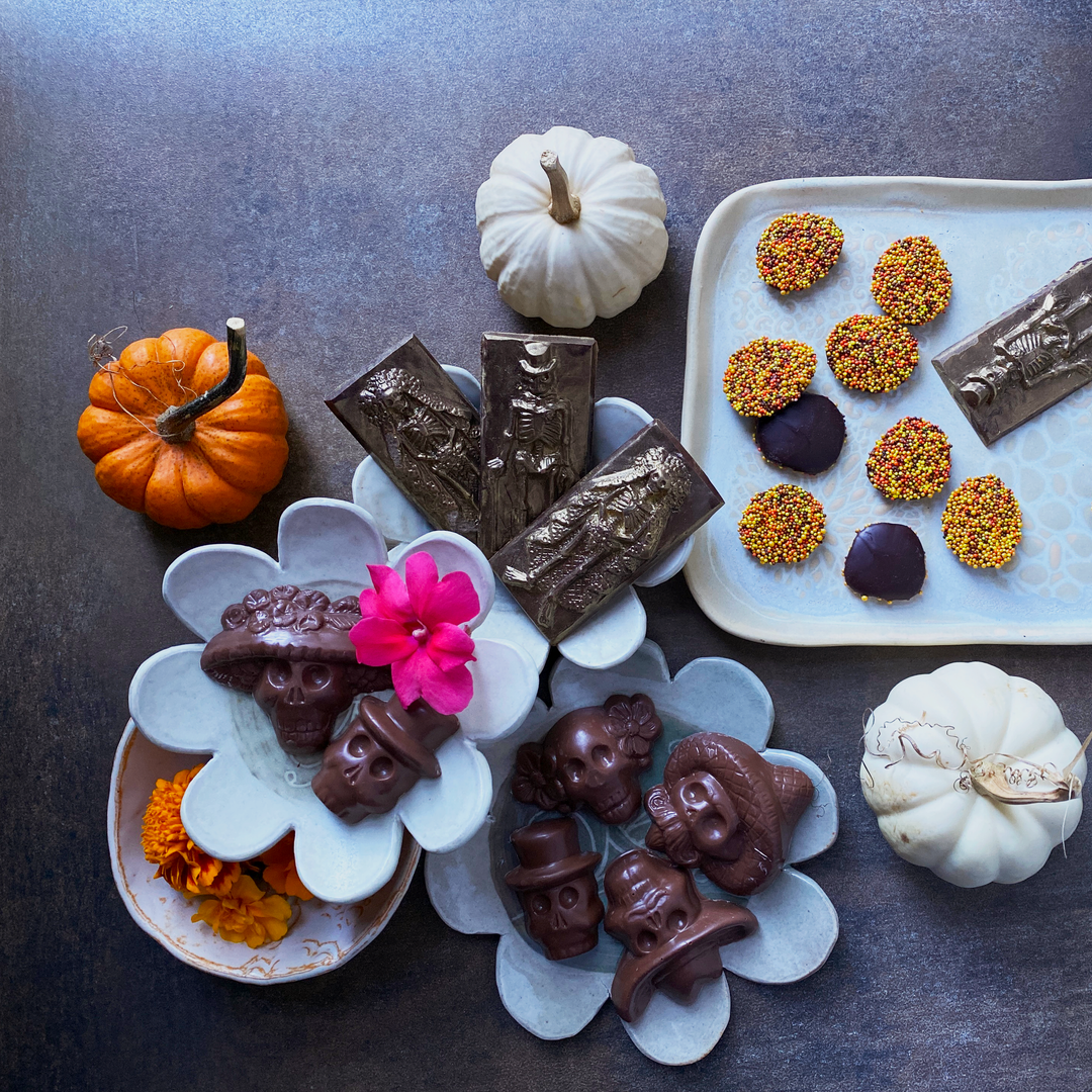 Assorted Day of the Dead themed chocolates, including skulls with hats, skeleton couples on gold bars, and round chocolates with orange and yellow sprinkles, arranged on a white background.