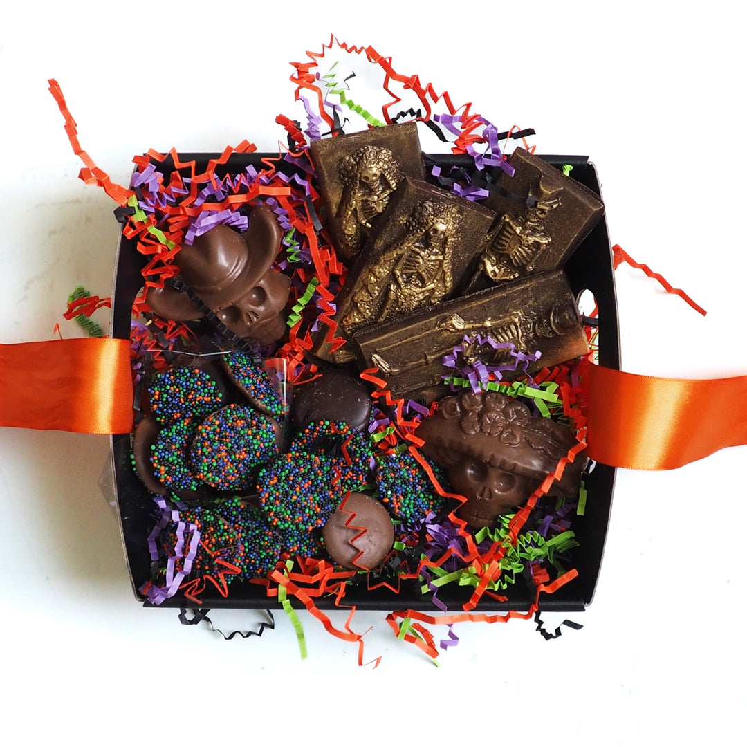 A Halloween Chocolate Party Bundle by Ragged Coast Chocolates, featuring skull-shaped chocolates, skeleton-embossed bars, and sprinkled dark chocolate coins nestled in orange, purple, and green paper shreds.