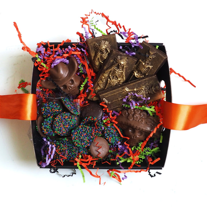 A Halloween Chocolate Party Bundle by Ragged Coast Chocolates, featuring skull-shaped chocolates, skeleton-embossed bars, and sprinkled dark chocolate coins nestled in orange, purple, and green paper shreds.