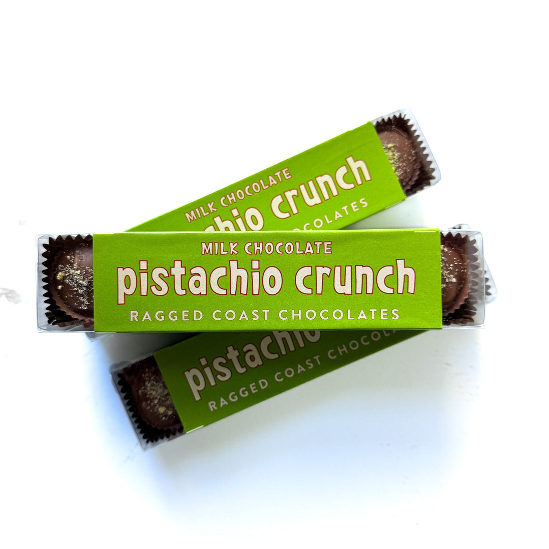 Two packages of Ragged Coast Chocolates Milk Chocolate Pistachio Crunch Cups, with green wrappers and text, are stacked diagonally against a white background.