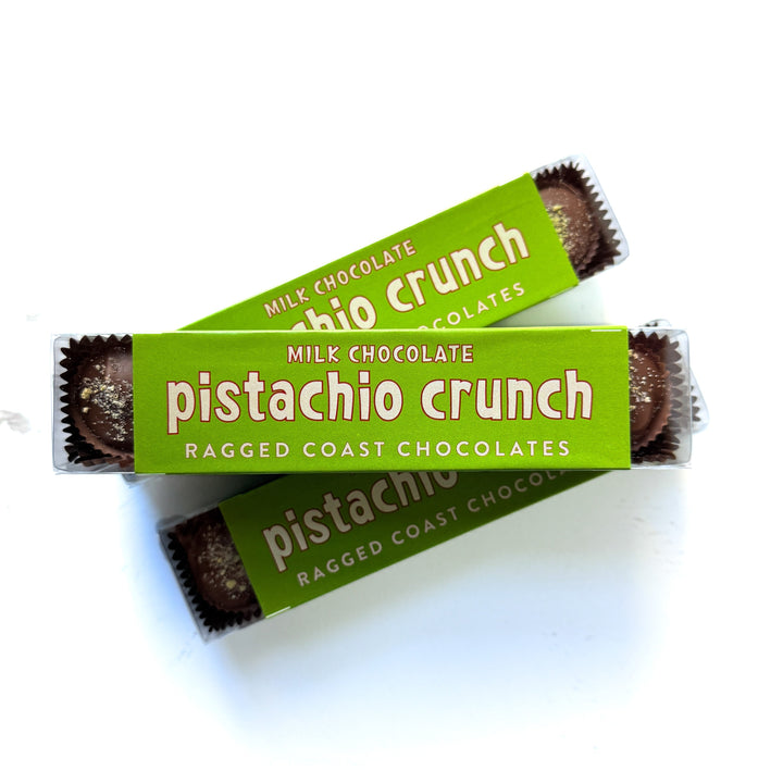Two packages of Ragged Coast Chocolates Milk Chocolate Pistachio Crunch Cups, with green wrappers and text, are stacked diagonally against a white background.