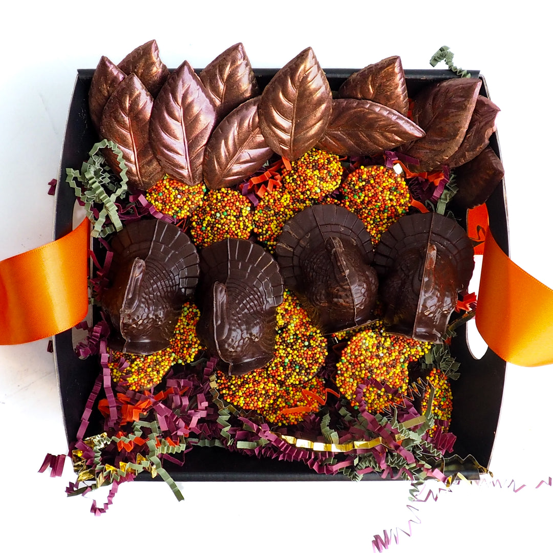 A Thanksgiving Chocolate Table Setting Bundle in a square box containing four chocolate turkeys and chocolate leaves, set on autumn nonpareils and shredded paper filler, with orange ribbons extending from the sides.