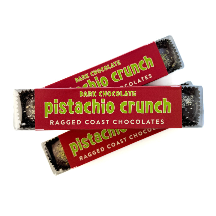 Two wrapped cups of Ragged Coast Chocolates Dark Chocolate Pistachio Crunch Cups, made with direct-trade chocolate, in red packaging with visible pieces of chocolate inside.