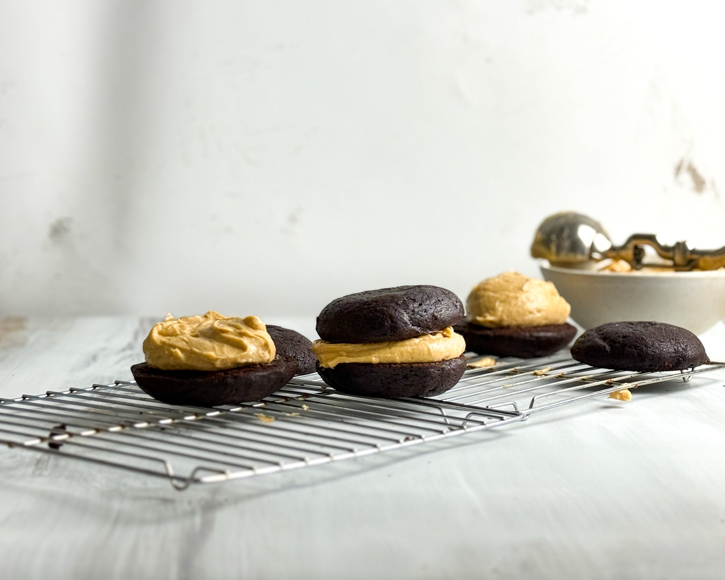 Chocolate whoopie pies with a creamy filling are cooling on a wire rack. In the background, a bowl and ice cream scoop sit on a light surface. The scene has a simple, airy feel.
