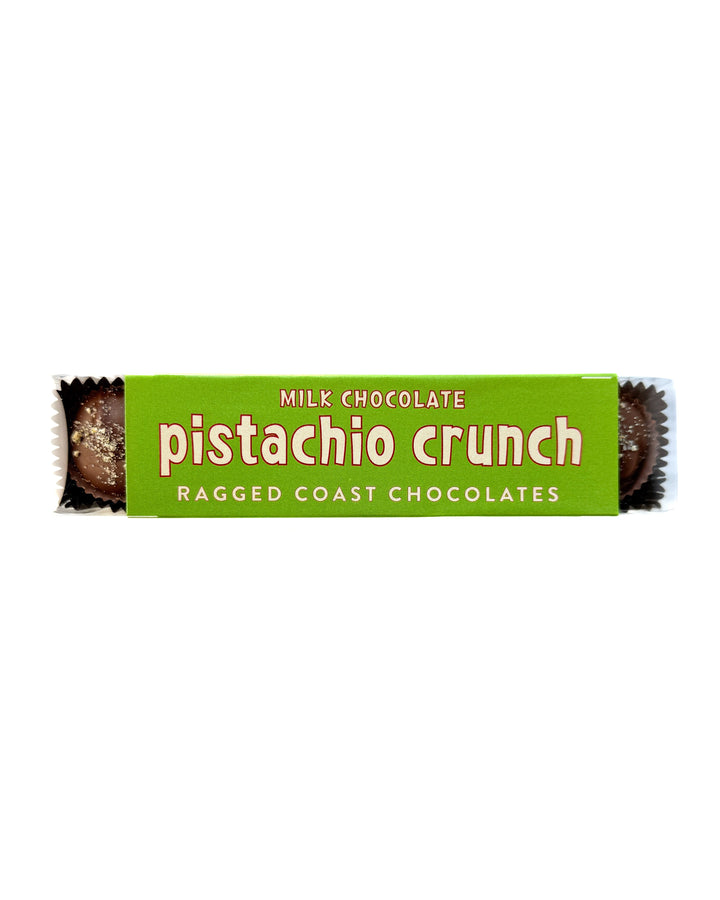 A green package labeled Milk Chocolate Pistachio Crunch Cups by Ragged Coast Chocolates, featuring direct-trade chocolate and handmade confections, with chocolate pieces partially visible at both ends.