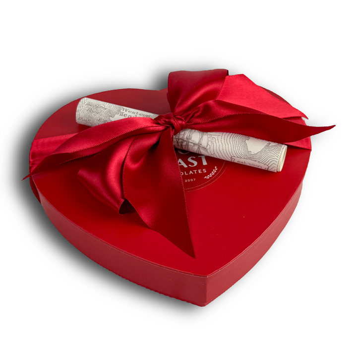 The Ragged Coast Chocolates Heart-Shaped Box - 18 Pieces features a red box with a satin bow and rolled parchment, perfect for presenting chocolate truffles, set against a white background.