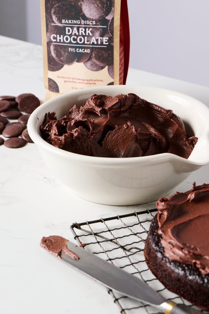A bowl of Ragged Coast Chocolates' Bittersweet Chocolate Baking Discs, 71% Cacao alongside baking chocolate and a partially frosted chocolate cake.