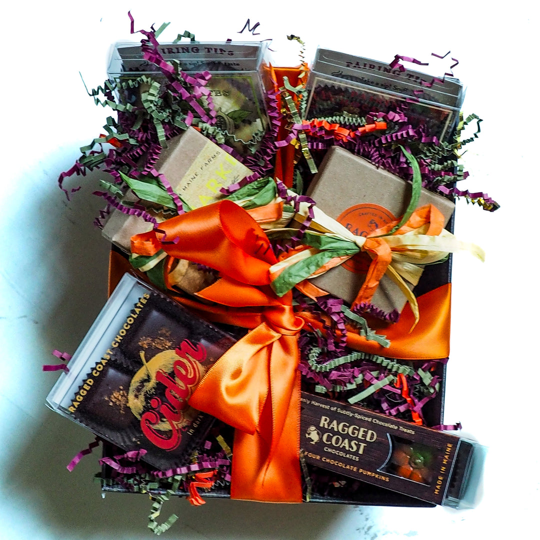 An Autumn Chocolate Sampler Gift Box by Ragged Coast Chocolates filled with assorted bars and boxes, accented with an orange ribbon and colorful shredded paper filler.