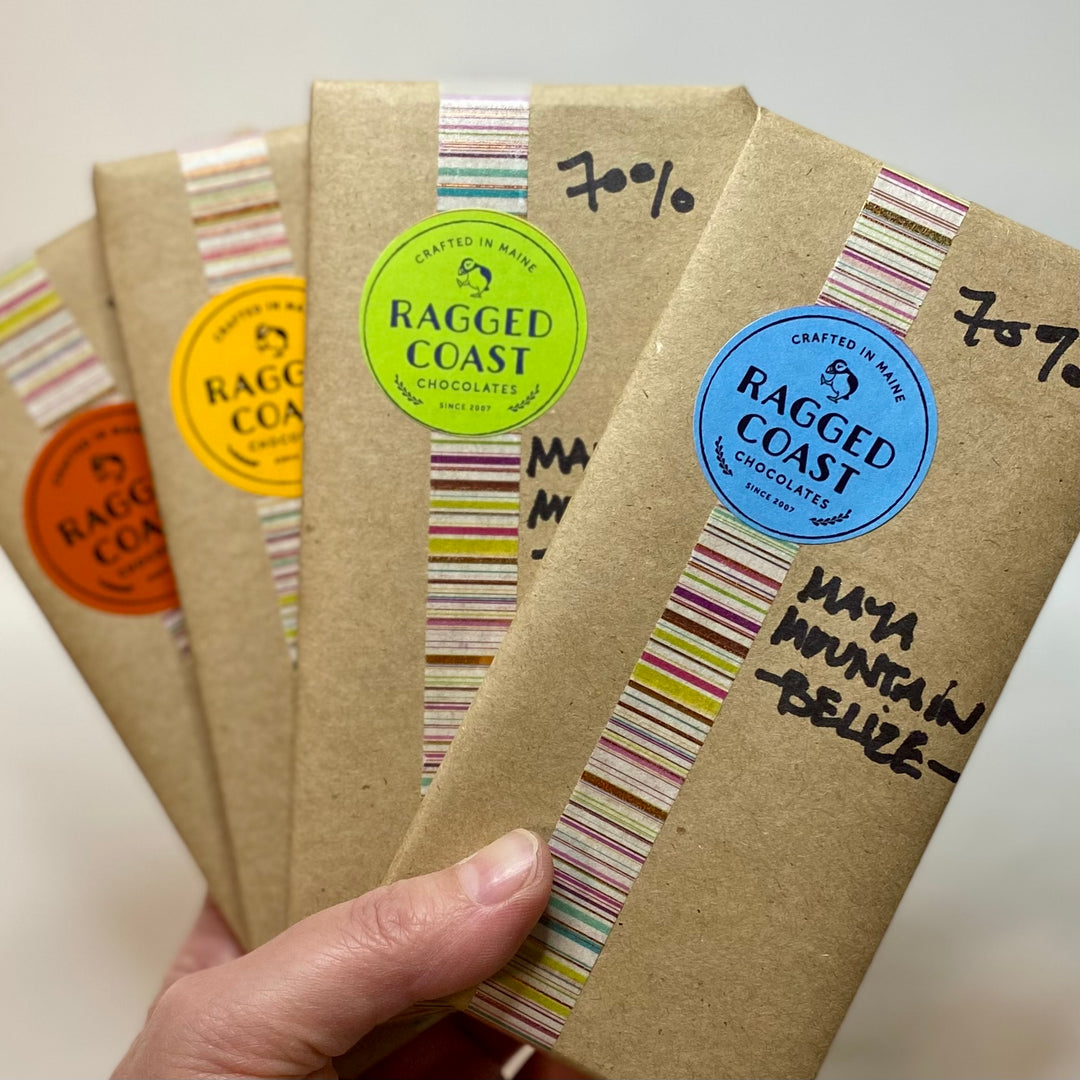 A hand holding a selection of ragged coast chocolates bars with various flavor labels, including the Single Origin Dark: Belize bar by raggedcoastchocolates.