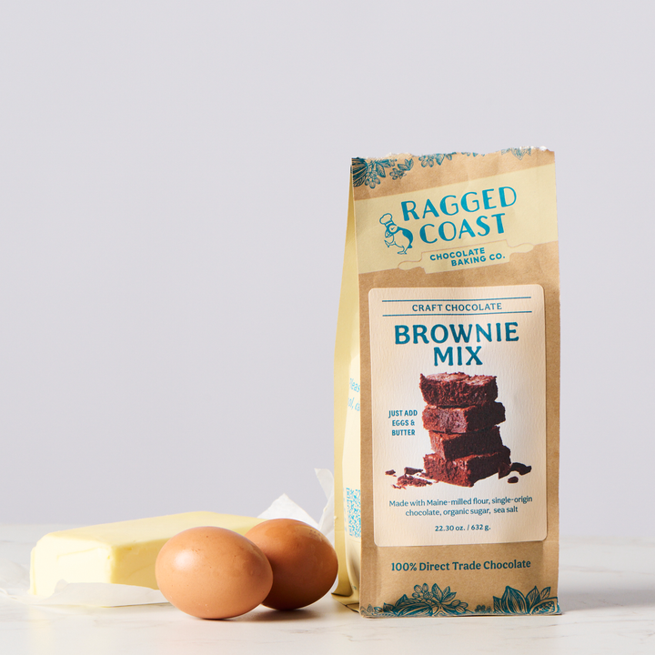 A package of Ragged Coast Chocolates Direct-Trade Cocoa Craft Chocolate Brownie Mix with eggs and a stick of butter on a kitchen counter.