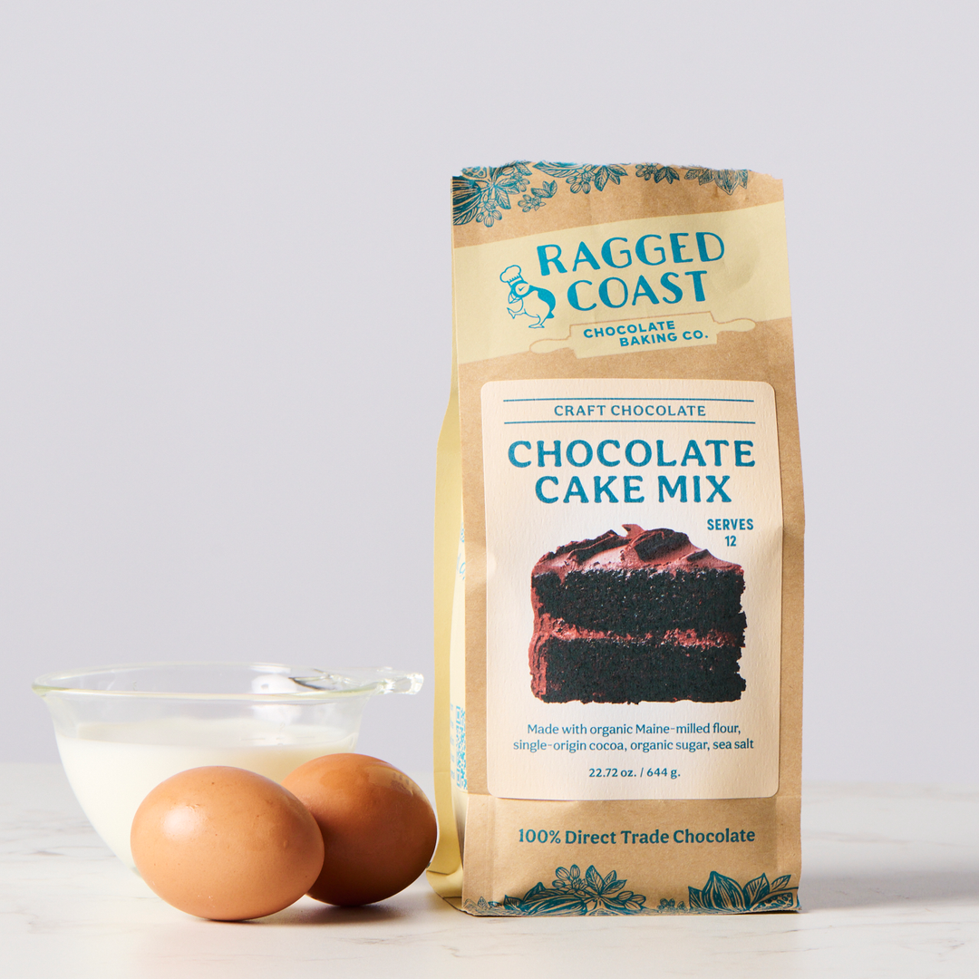 A package of Ragged Coast Chocolates chocolate cake mix made with direct-trade cocoa and organic sugar, with a clear bowl of eggs on a kitchen counter.