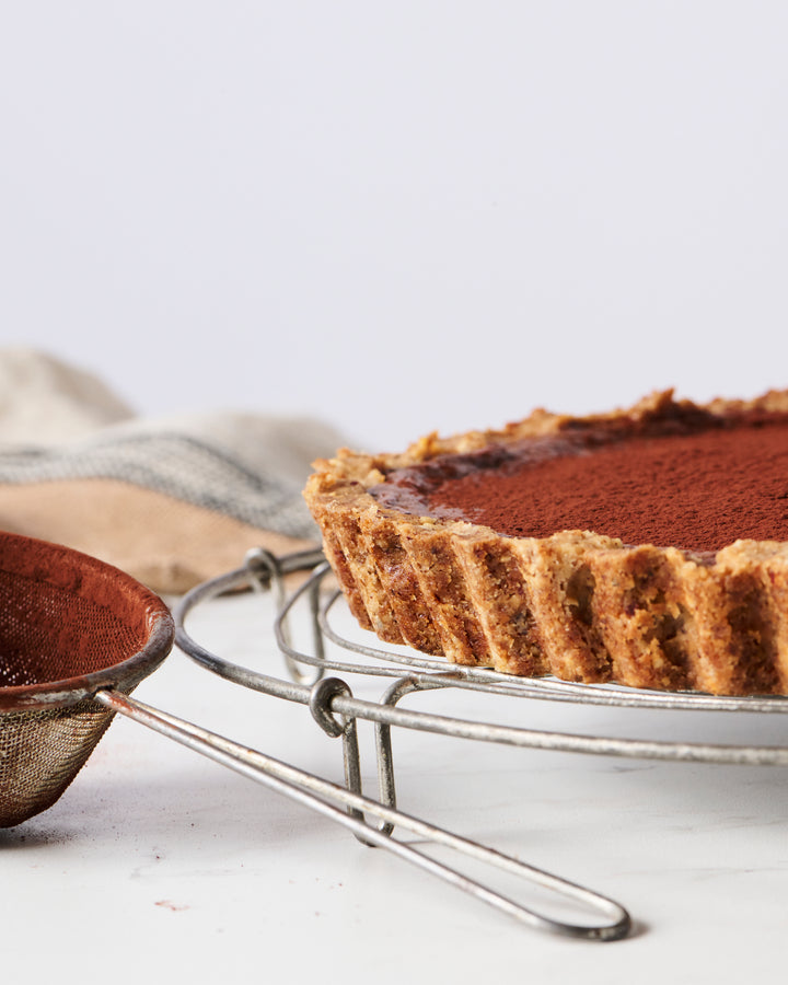 A close-up of a freshly baked pie with a dusting of cocoa powder on top, enhanced with Ragged Coast Chocolates' Direct Trade Ecuadorian 40% Caramelized Milk Chocolate ganache, placed on a wire cooling rack.