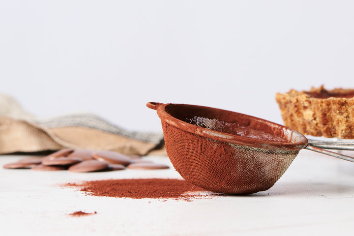 Clay bowl with Ragged Coast Chocolates' Exceptionally Dark Cocoa Powder with high cocoa butter content and a mesh sieve on a white surface, chocolate pieces and a pie in the background.