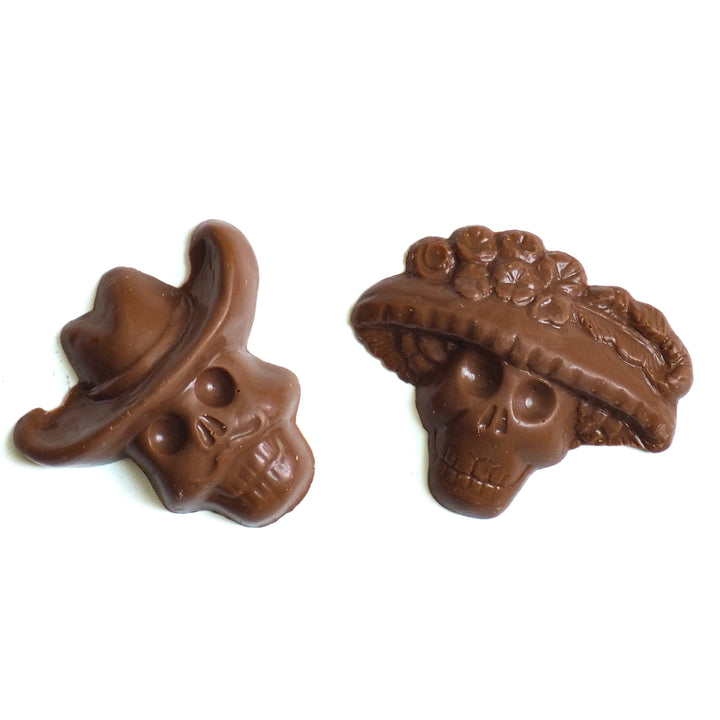 Two dark chocolate Day of the Dead skulls from the Halloween Chocolate Party Bundle, one shaped like a cowboy wearing a hat and the other like a figure wearing a floral hat.