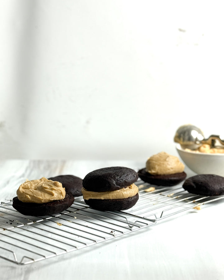 Four whoopie pies with creamy filling, made from Ragged Coast Chocolates' Dark Chocolate and Peanut Butter Whoopie Pie Mix, are arranged on a wire rack. In the background are a bowl of extra filling and an ice cream scoop on a white minimalist surface.
