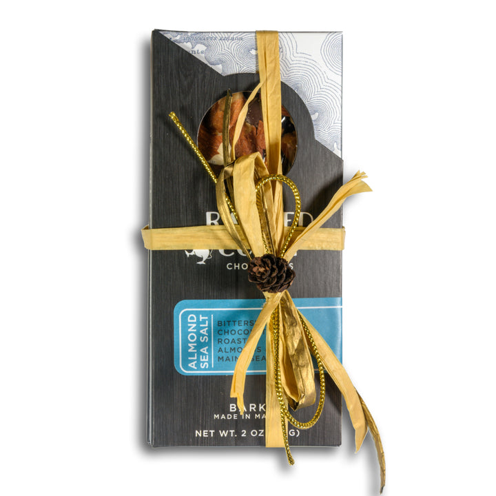A Ragged Coast Chocolates Chocolate Bark Bundle: Holiday Edition with organic roasted almond bits wrapped as a gift and adorned with a yellow ribbon and a pine cone decoration.