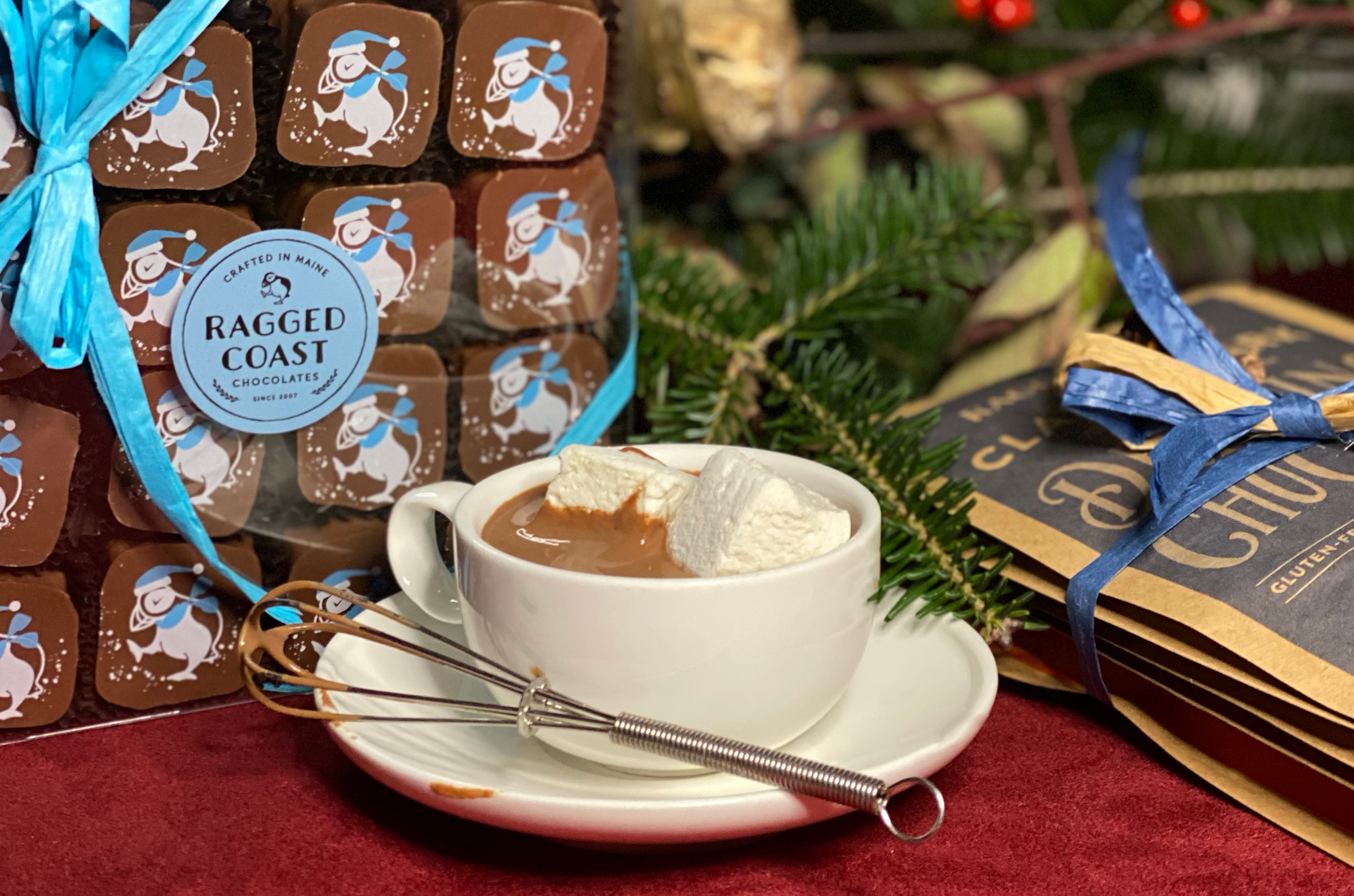 A cup of hot chocolate with marshmallows, surrounded by chocolate bars and holiday decorations.