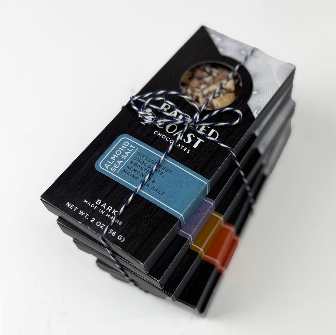 A stack of black Ragged Coast Chocolates boxes, tied with string. The top box has a window with mixed nuts. A blue label reads Almond Maine sea salt dark chocolate bark. Colorful labels mark some Chocolate Bark Bundle boxes as vegan and gluten free.