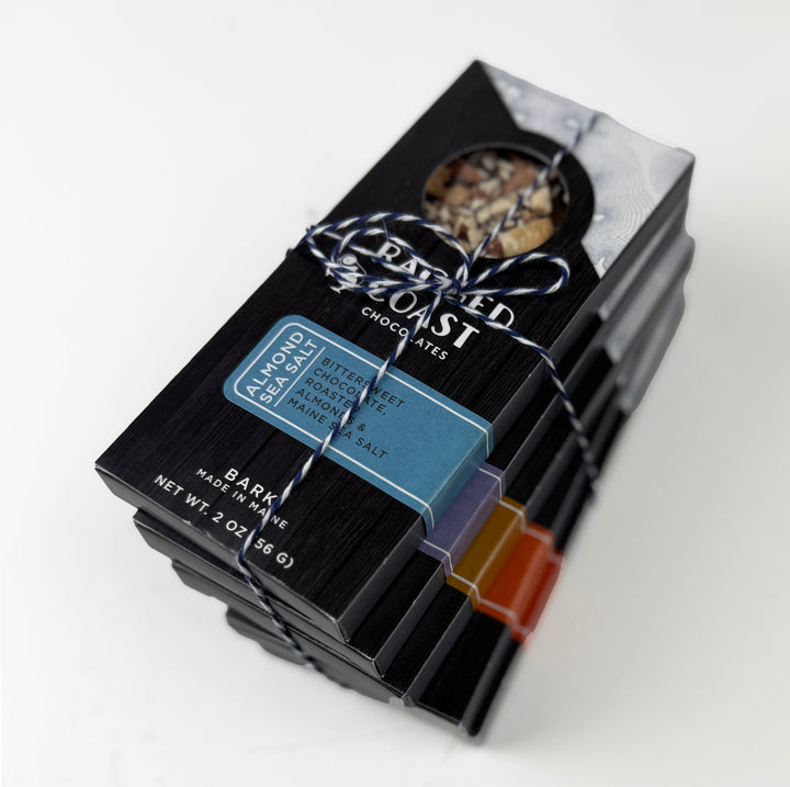 A stack of black Ragged Coast Chocolates boxes, tied with string. The top box has a window with mixed nuts. A blue label reads Almond Maine sea salt dark chocolate bark. Colorful labels mark some Chocolate Bark Bundle boxes as vegan and gluten free.