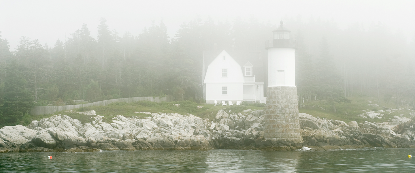 A lighthouse and adjacent building shrouded in mist along a rocky coastline.