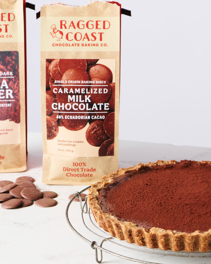 A freshly baked chocolate tart with a dusting of cocoa next to packages of Ragged Coast Chocolates Direct Trade Ecuadorian 40% Caramelized Milk Chocolate baking discs perfect for ganaches and frostings.
