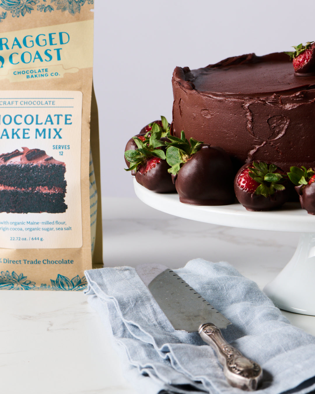 A chocolate cake topped with strawberries next to a Ragged Coast Chocolates Baking Mix Gift Box and a cake server on a cloth.
