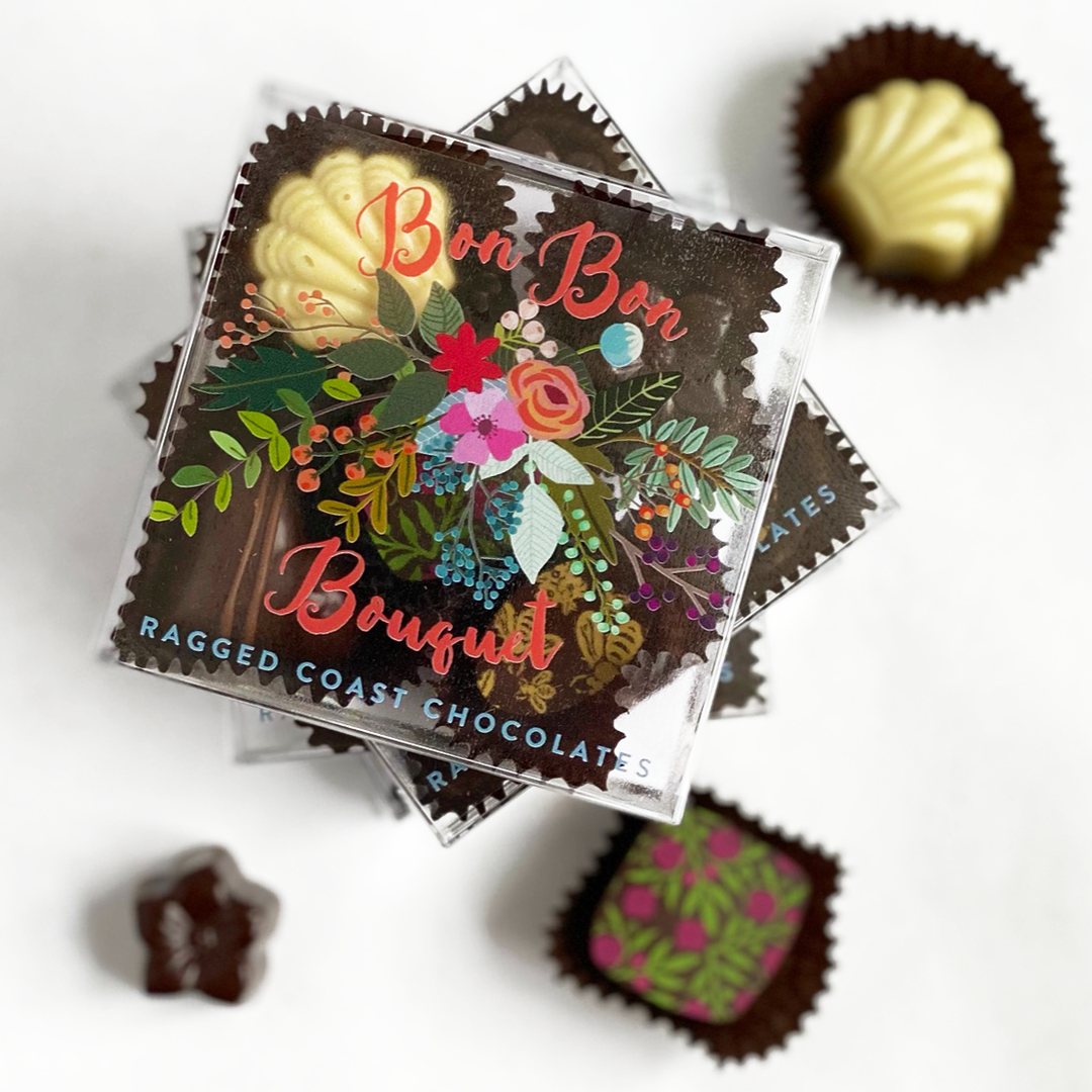 Top view of a stack of Mother's Day chocolates labeled "The Gardener Bundles" by Ragged Coast Chocolates, with two unwrapped chocolates beside them.