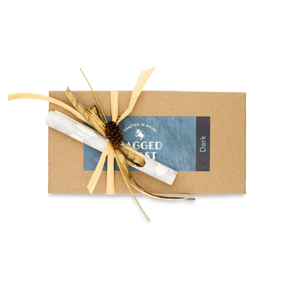 A brown rectangular gift box, made from recyclable paperboard and tied with a raffia ribbon, is decorated with a small pinecone and features a blue label with partially visible text. Inside, enjoy the delightful Dark Chocolate Truffle Assortment by Ragged Coast Chocolates.