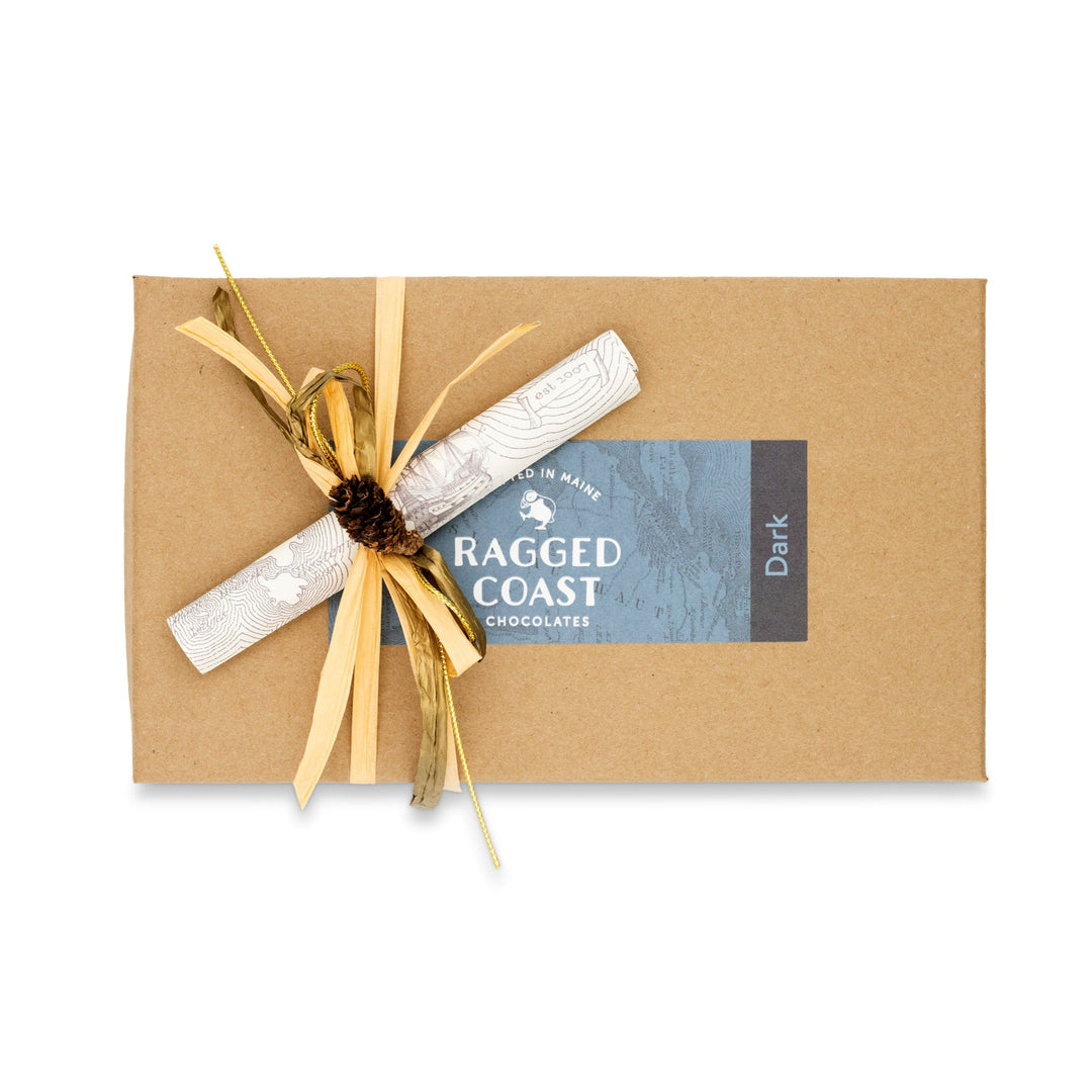 The Dark Chocolate Truffle Assortment from Ragged Coast Chocolates comes in a brown box adorned with a blue label reading "Dark," tied with ribbon and a pine cone. Inside, delectable dark chocolate truffles are nestled within recyclable paperboard boxes, combining indulgence and sustainability.