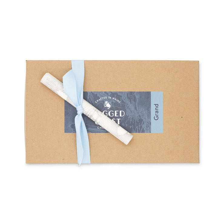 A brown paper gift box tied with a blue ribbon and a decorative tag, containing Ragged Coast Chocolates' Grand Assortment of Milk and Dark Chocolate Truffles.