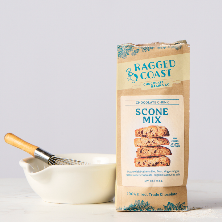 A bag of Ragged Coast Chocolates Chocolate Chunk Scone Mix placed next to a mixing bowl and a whisk on a kitchen countertop.