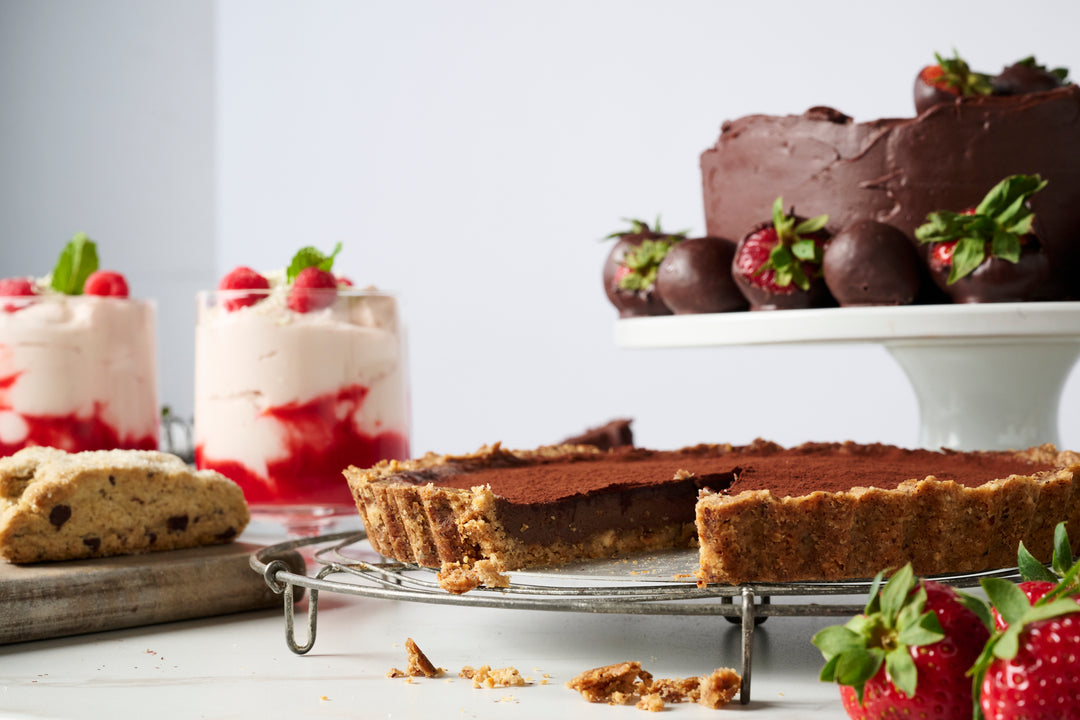 An assortment of desserts including a Ragged Coast Chocolates Direct Trade Ecuadorian 40% Caramelized Milk Chocolate tart, strawberry parfaits, and a biscotti on a white background.