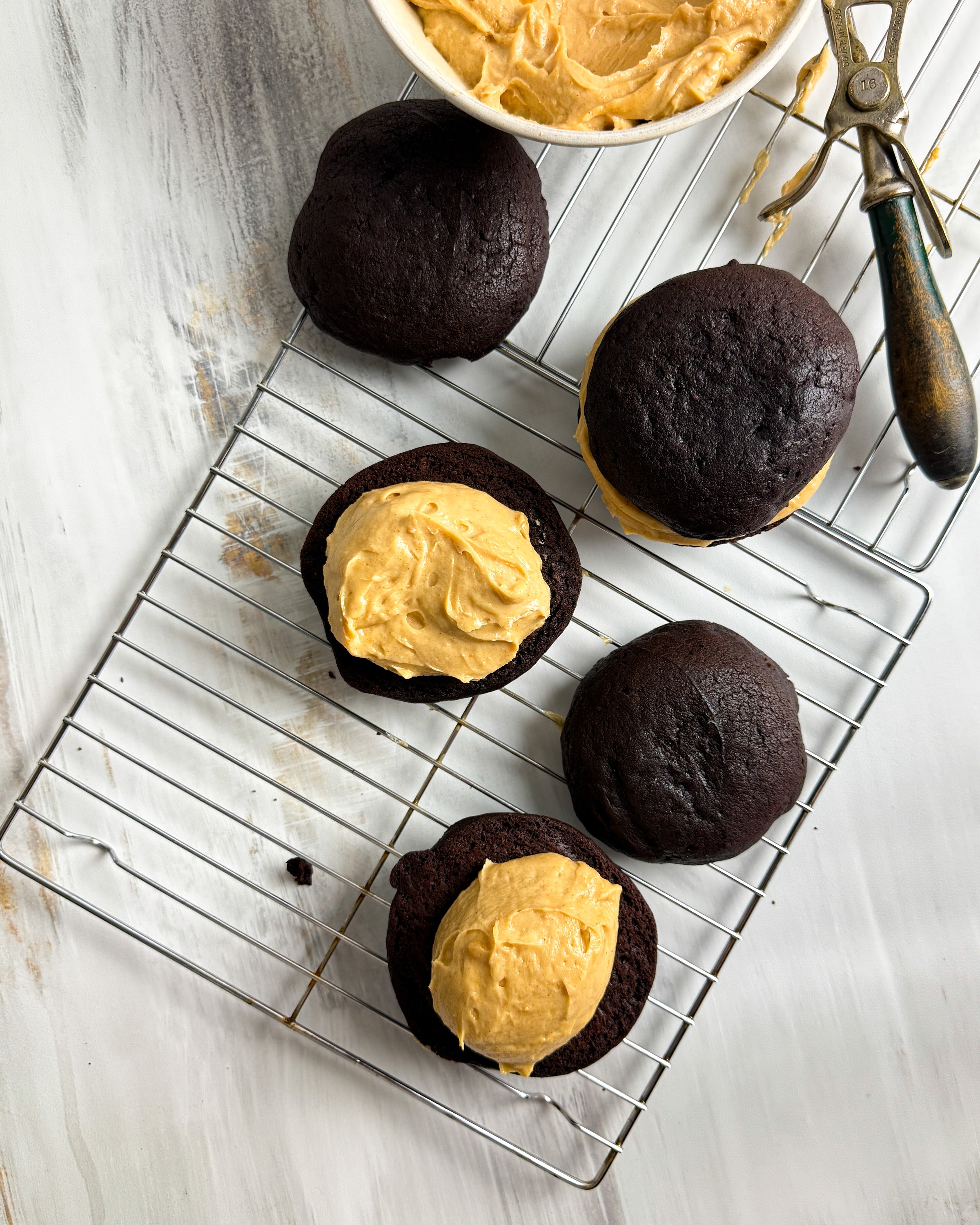 whoopie pies with peanut butter filling on a cooling rack with a bowl of peanut butter filling in the background.