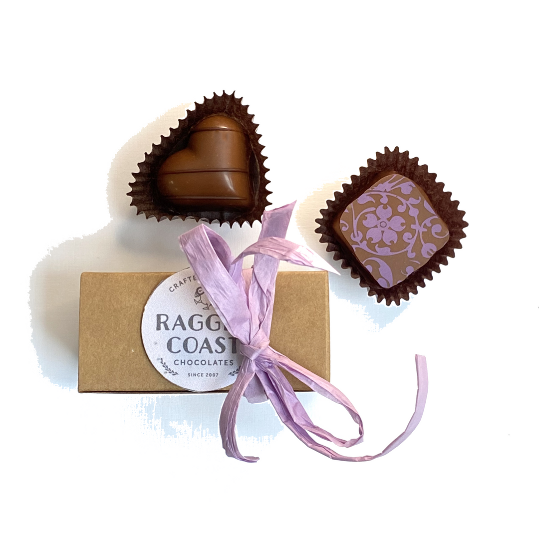 A selection of fine chocolates from the Ragged Coast Chocolates Grand Assortment Collection next to a gift-wrapped box with a purple ribbon.