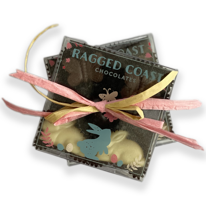 Two stacked Bunny & Butterfly Bonbon boxes from Ragged Coast Chocolates are tied with a pink and yellow ribbon, featuring pastel bunnies and eggs on a dark background, ideal for spring celebrations.