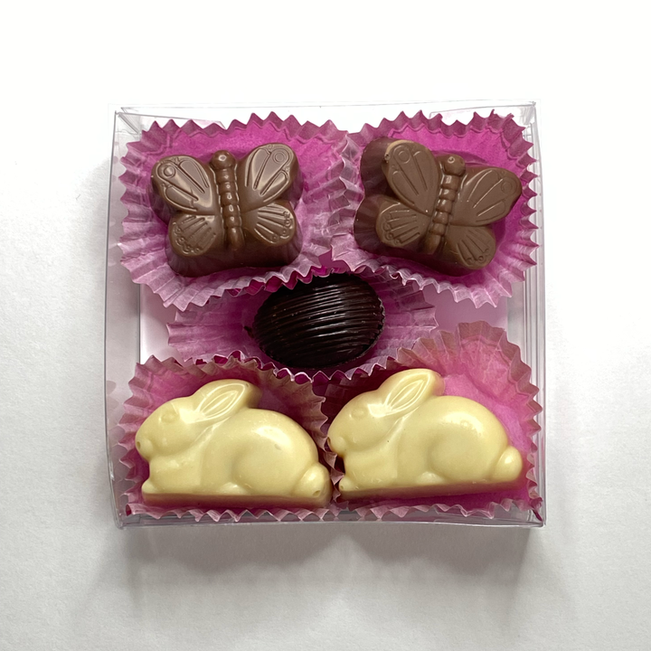 The Ragged Coast Chocolates Bunny & Butterfly Bonbons come in a clear box with two butterfly-shaped, one oval, and two rabbit-shaped chocolates nestled in pink paper cups, ideal for spring festivities.