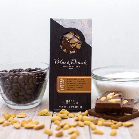 A package of Ragged Coast Chocolates' gluten-free Chocolate Coffee Peanut Bark, accompanied by a bowl of coffee beans, a glass of milk, and pieces of the chocolate bar with organic peanuts.